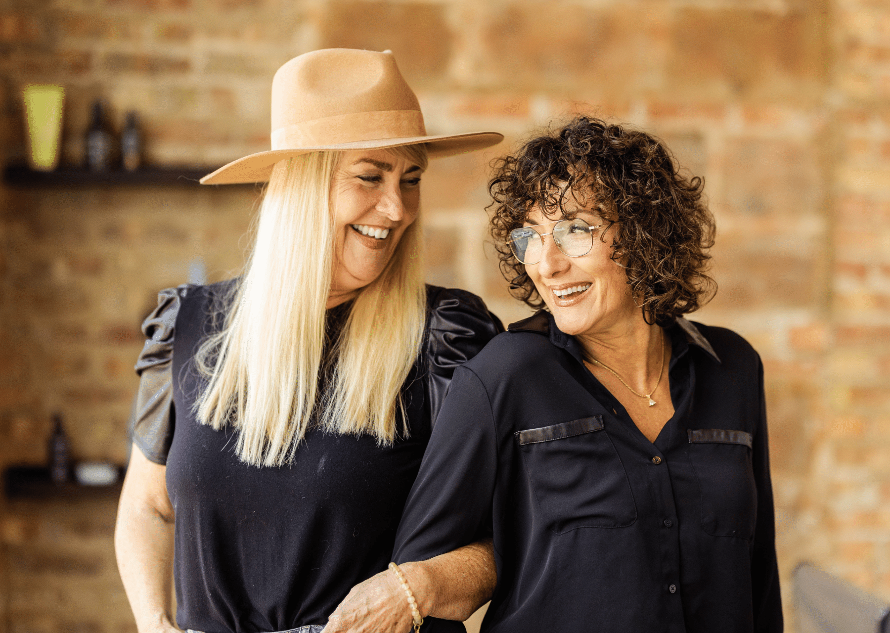 A long-haired, blonde woman in a wide-brimmed hat linking arms with a brunette, curly-haired wmoman, both smiling and mid-laughter